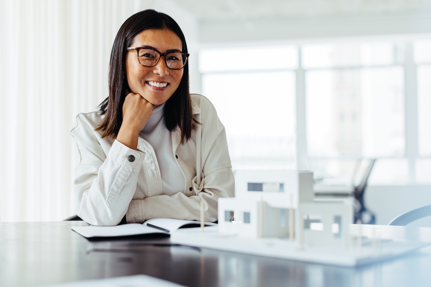 Female architect smiling at the camera while sitting with a 3D house model in an office. Creative business woman working on a building design project.