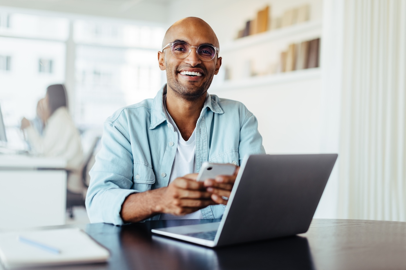 Business man smiling at the camera while holding a mobile phone. Young male designer sitting in an office with a laptop.